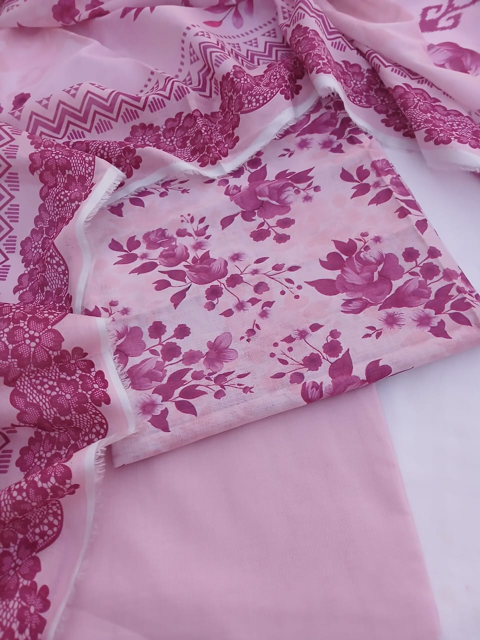 Flatlay view of an unstitched 3-piece printed lawn suit featuring a pastel pink floral shirt, solid trouser, and matching dupatta fabric.